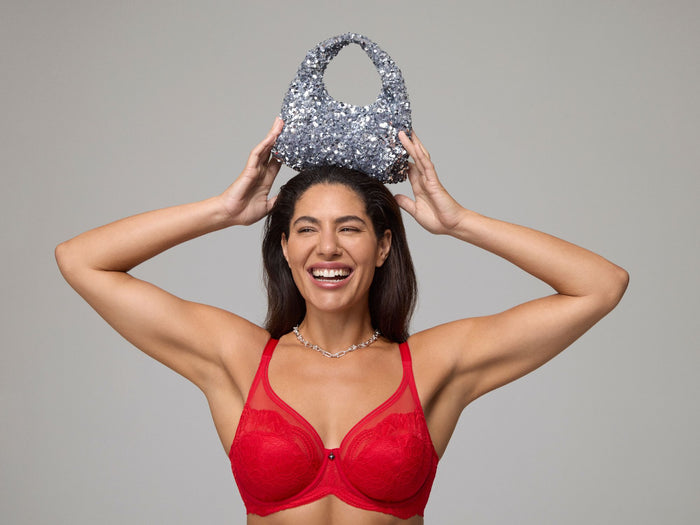 Woman wearing a red bralette holding a sparkly handbag above her head against a gray background