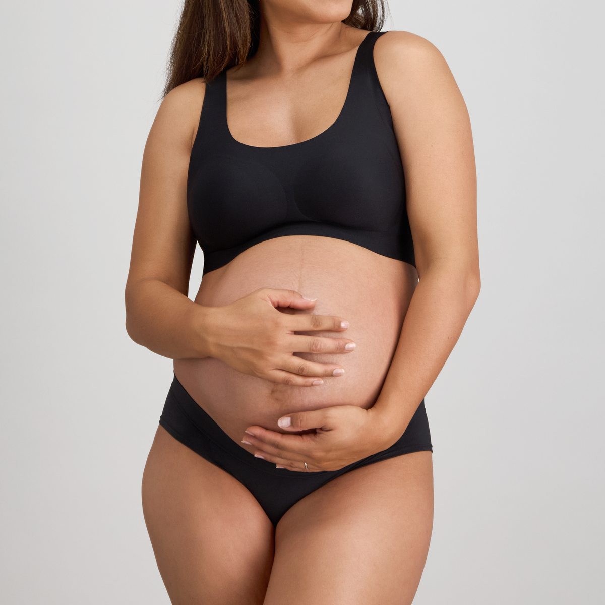 Pregnant woman wearing a black maternity bra and underwear set on a light gray background