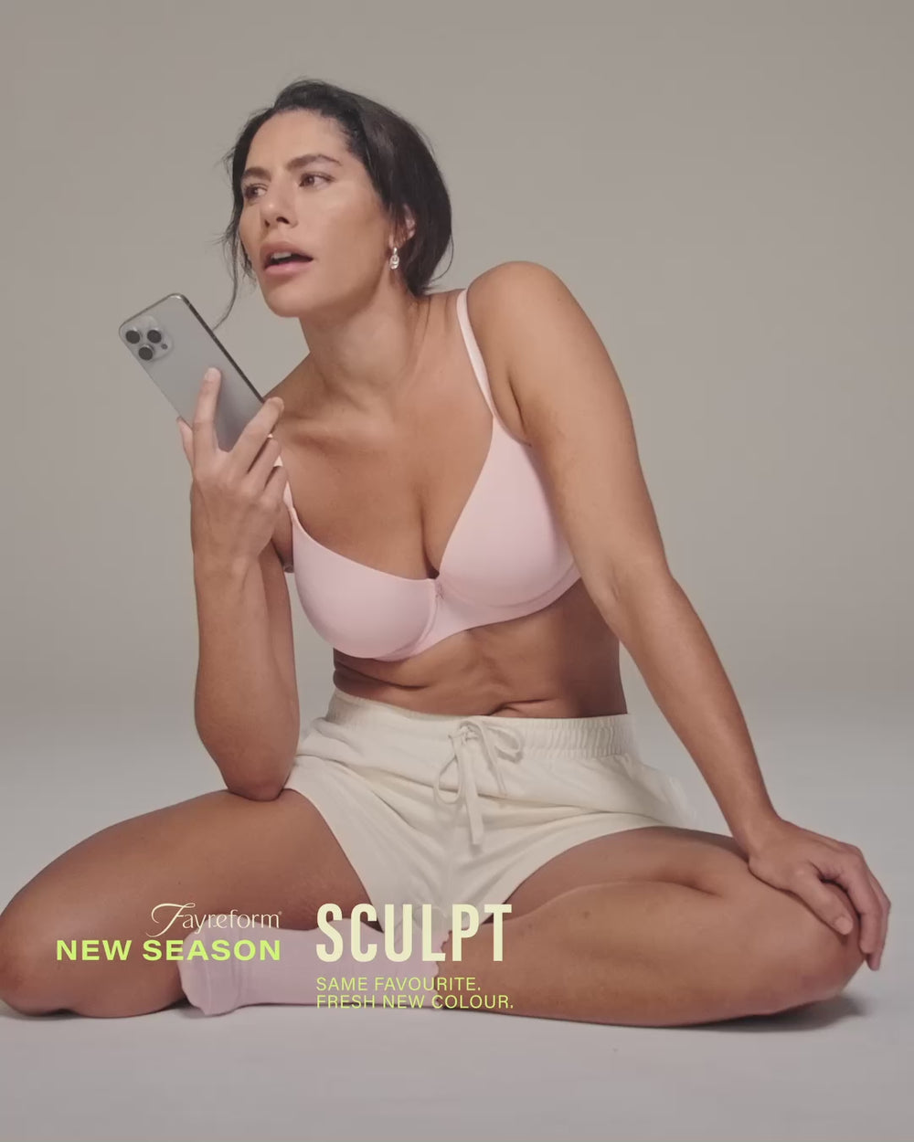 model-poses-holding-phone-wearing-light-pink-bra-and-white-shorts