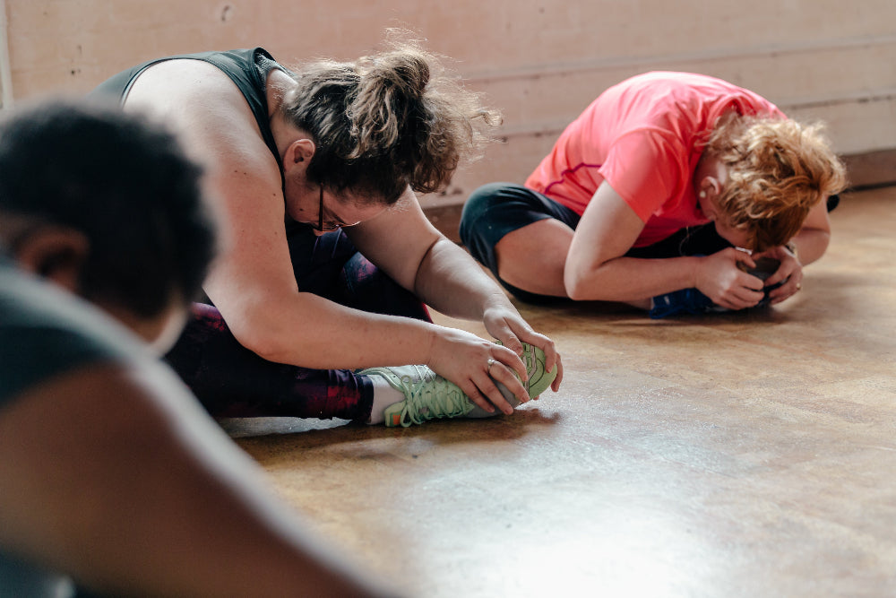 Two people stretching on a wooden floor in a gym or studio, with one in a black tank top and the other in a coral-pink t-shirt.